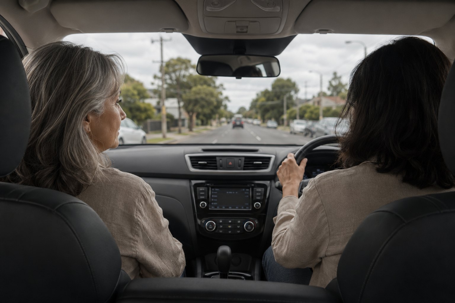 Two women in the front seats of a car on a suburban street, one looking out of the side window.